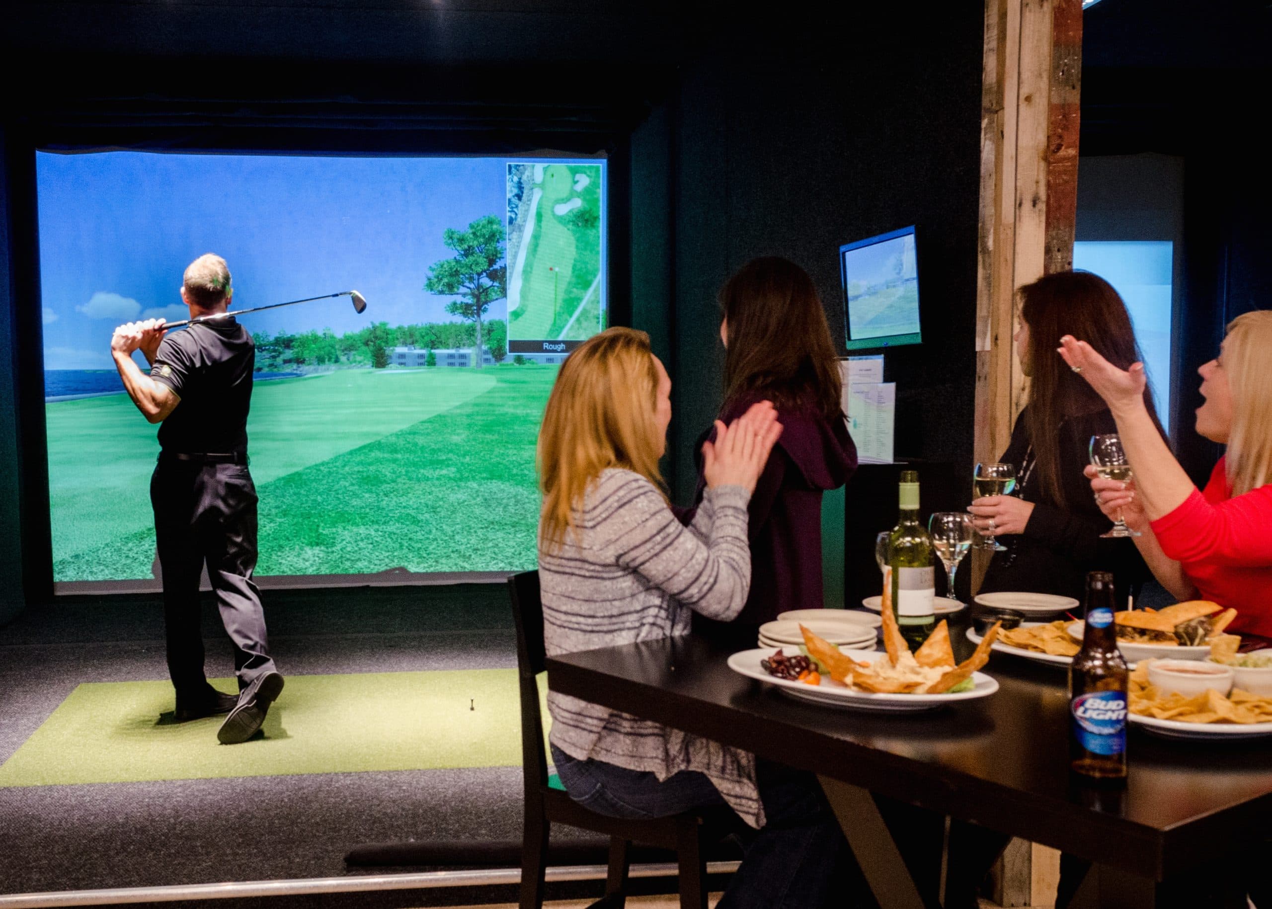 Friends cheering at a high-top table while a golfer tees off on the simulator